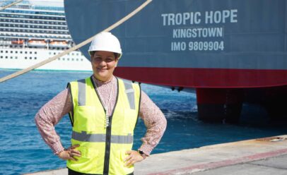 Keyla stands in front of the stern of the Tropic Hope, wearing a hard hat and reflective vest.