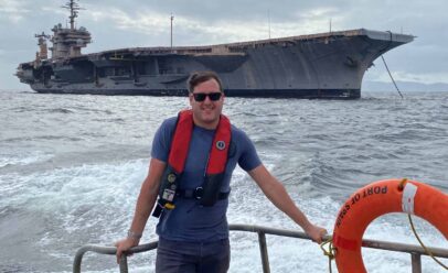 Michael Loomis leans on the rail of a Foss tug. The USS Kitty Hawk behind him.