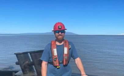 Eric Nyce smiles in hard hat and PFD. A vast stretch of ocean behind him.